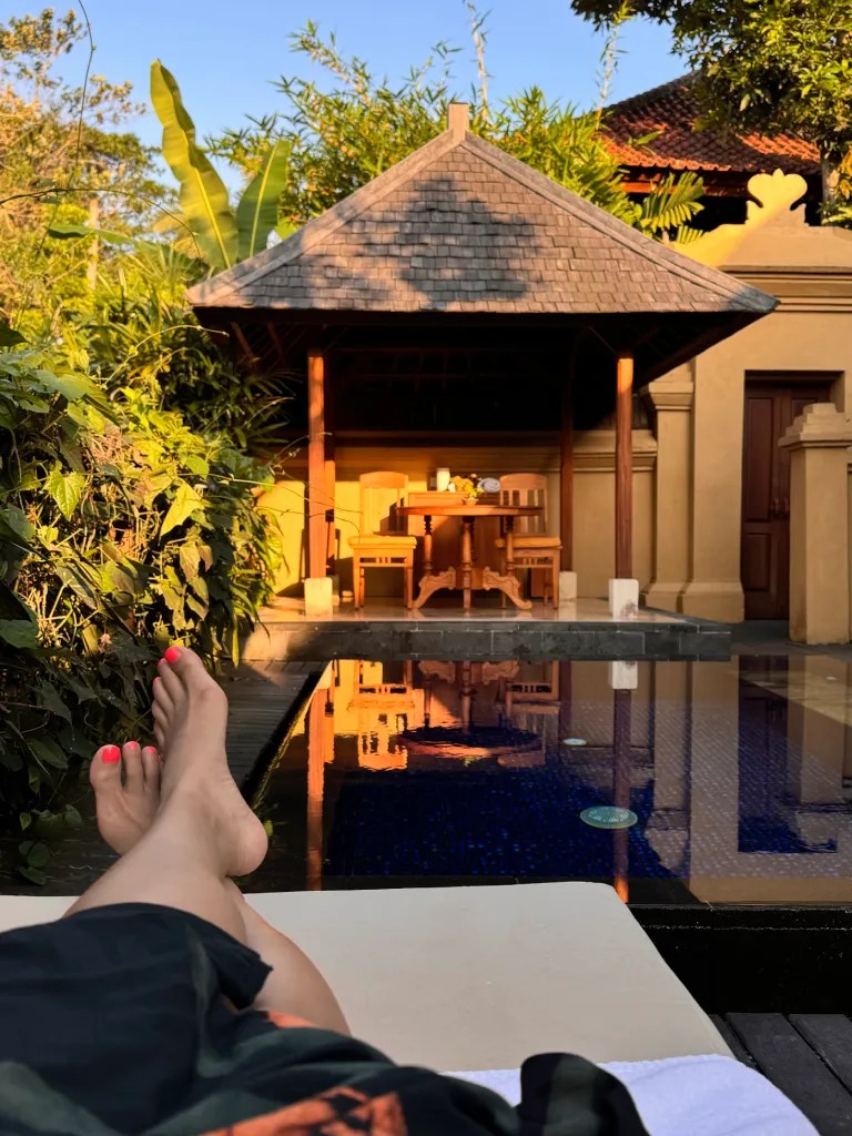 A person's feet with pink toenails resting on a lounge chair, with a scenic view of a thatched gazebo and a serene pool surrounded by lush greenery in Bali.