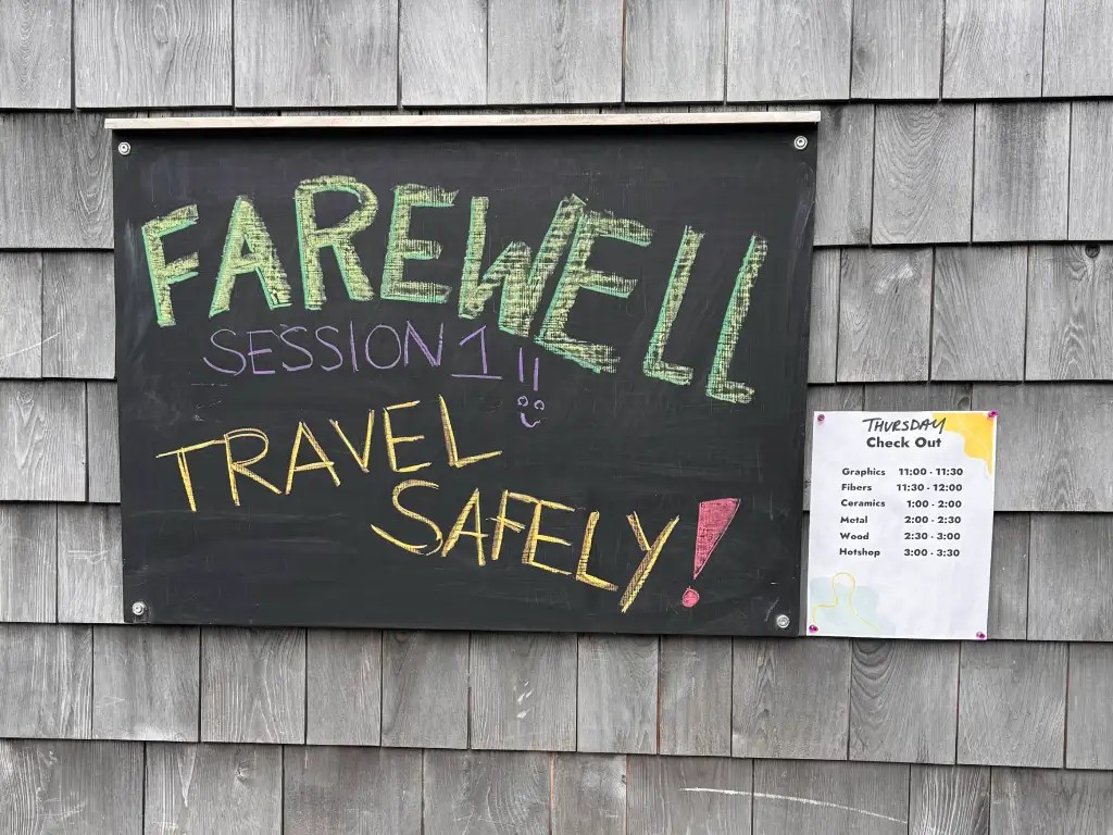 A chalkboard sign displaying a farewell message for Session 1, with colorful text and a friendly reminder to travel safely, alongside a schedule for check-out times for various craft studios.