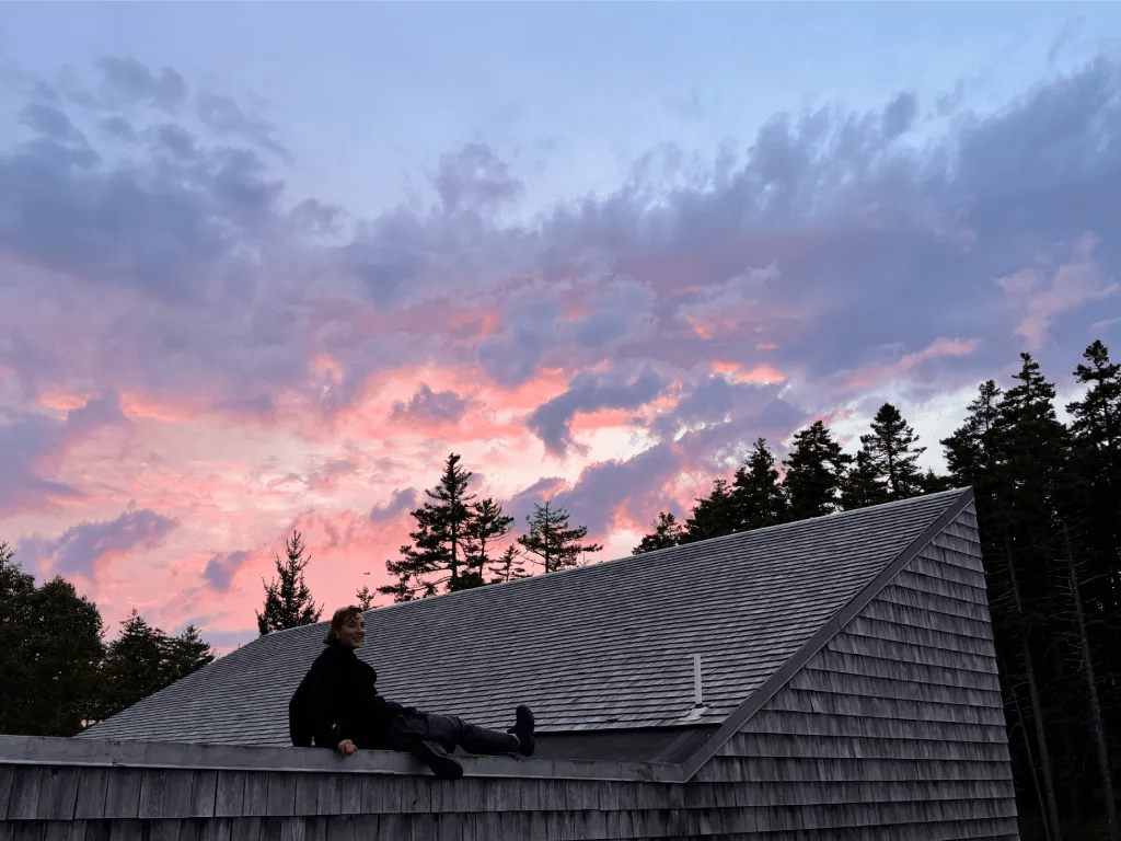 A person sitting on the roof of a studio at Haystack, looking at a colorful sunset sky filled with pink and purple hues, with trees in the background.