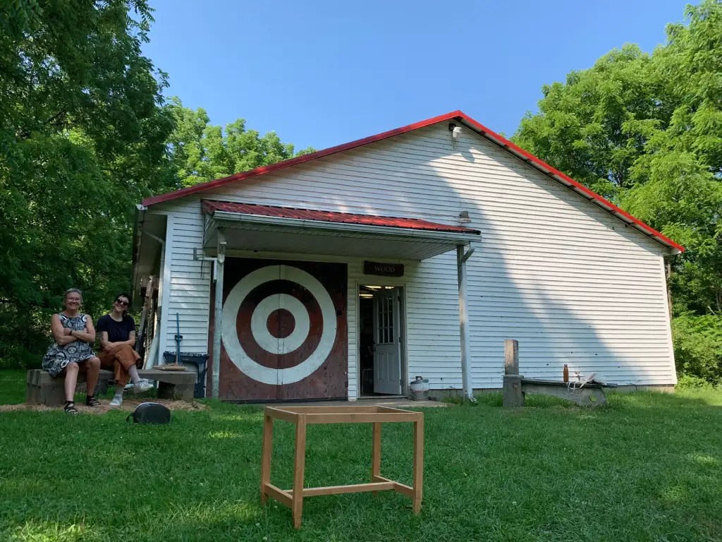 Two women sitting outside a craft school building with a target design on the door, with a finished wooden bench frame  on the grass in the foreground.