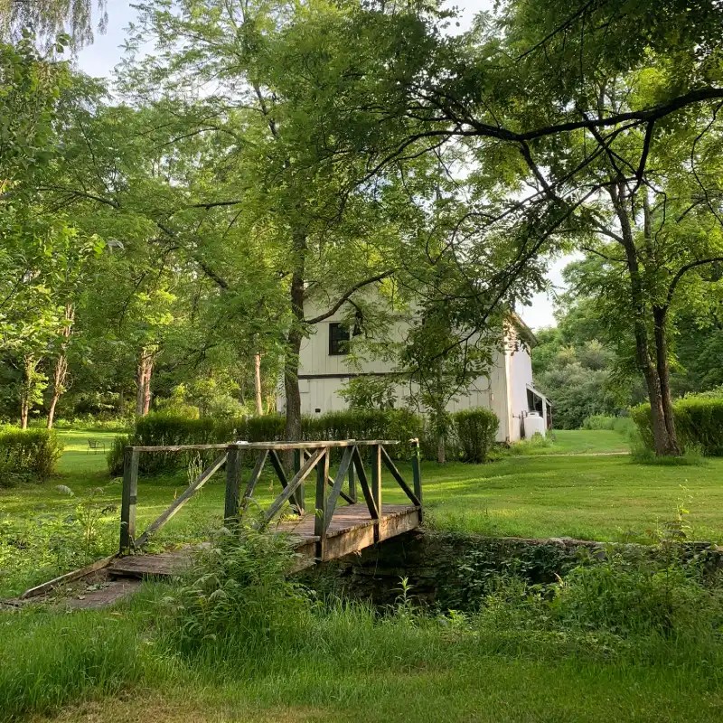 A wooden bridge crossing over a small stream, surrounded by lush greenery and trees, leading to a white building in a serene outdoor setting.