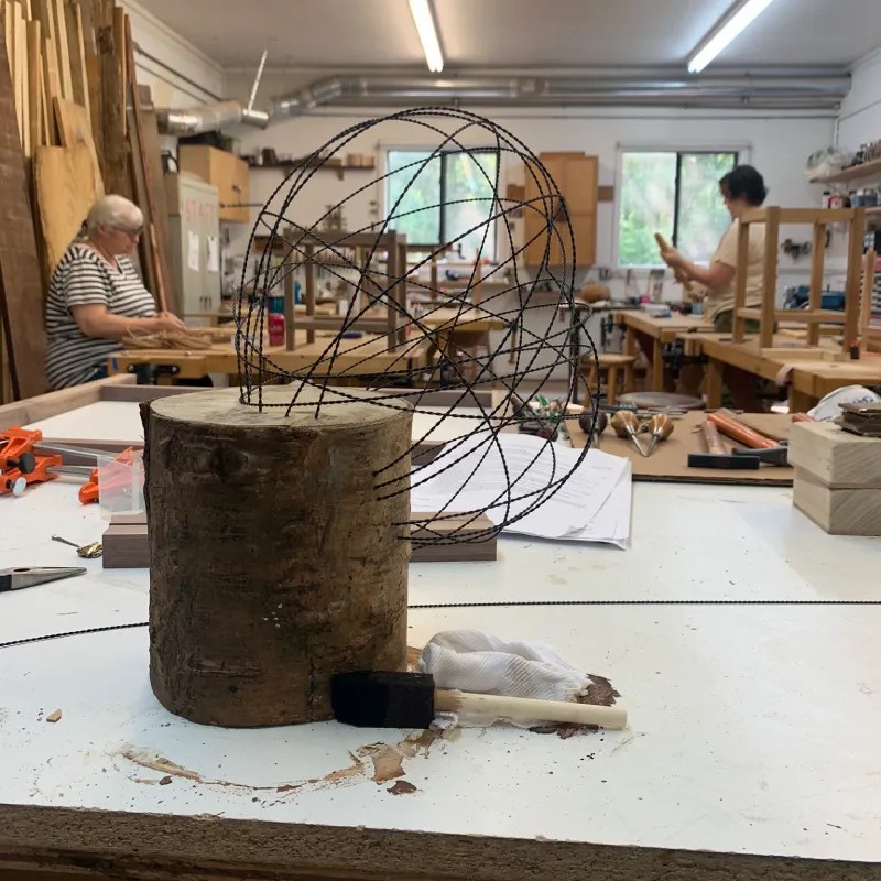 A wooden base supporting a wire sculpture in a craft studio, with various woodworking tools and materials visible in the background.