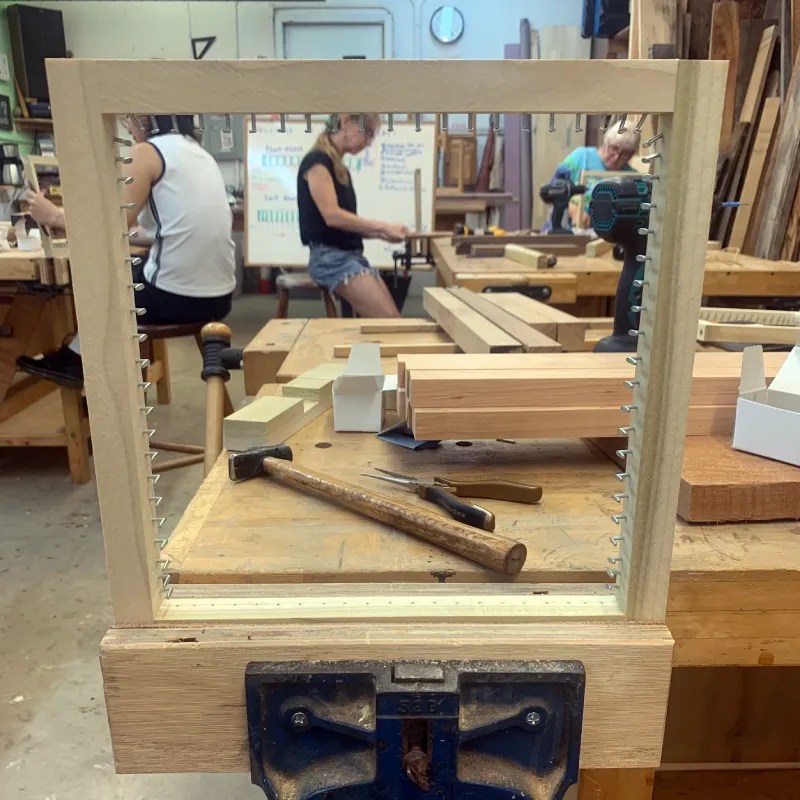 A wooden frame being assembled in a workshop, with various tools and wooden materials visible on the workbench, and workshop participants working in the background.