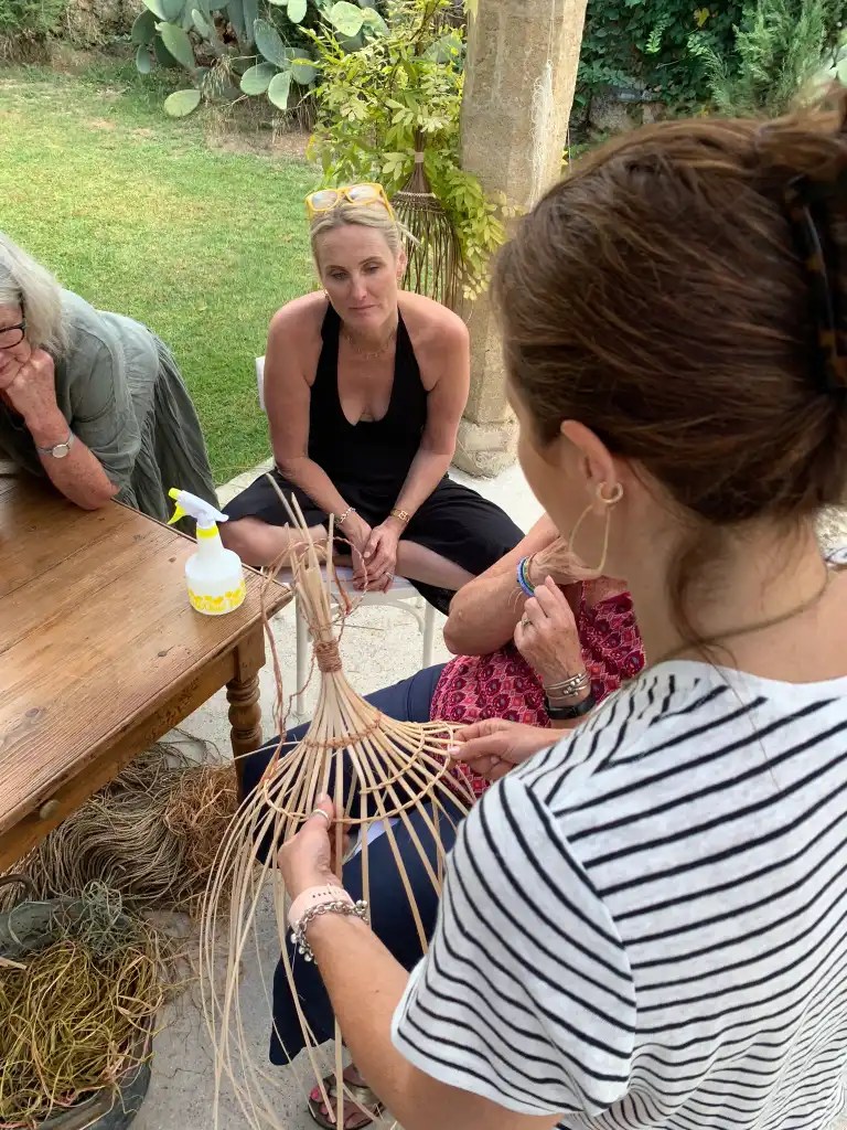A group of women gathered around a table in an outdoor setting, observing a woman demonstrating basket weaving techniques using natural materials.