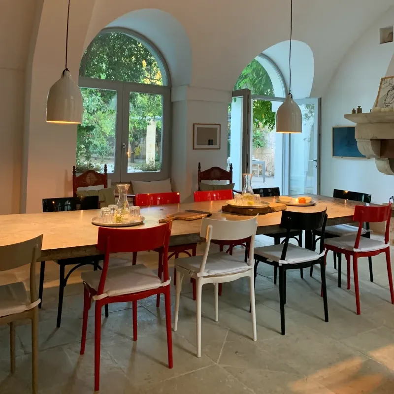 A spacious dining area with a long table surrounded by colorful chairs in a villa, featuring large windows that bring in natural light.