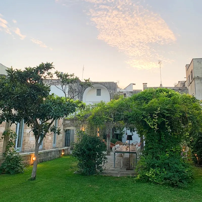 A picturesque villa in Muro Leccese, Italy, surrounded by lush greenery and an outdoor seating area under a vine-covered pergola, set against a pastel sky.