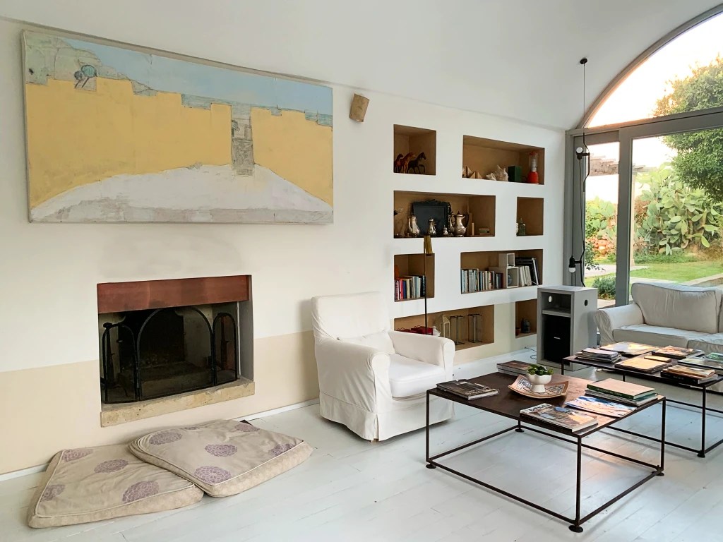 A cozy living room featuring a white sofa, an iron coffee table with books, and a colorful painting hanging above the fireplace.