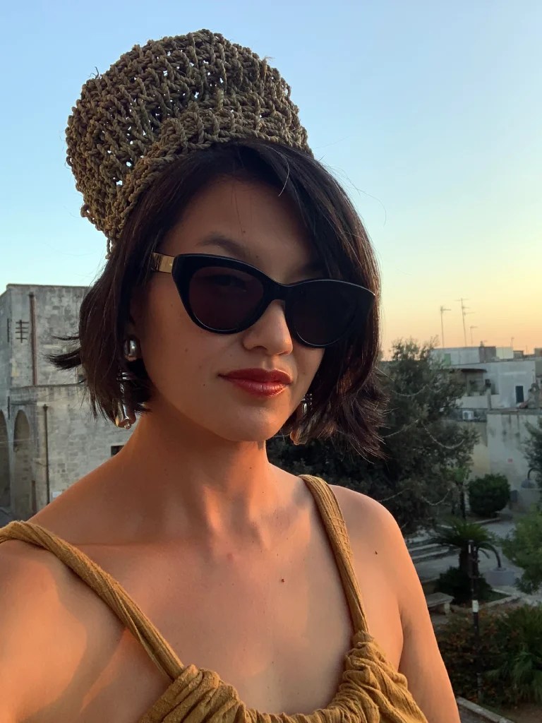 A person wearing a handwoven basket as a hat, dark sunglasses, and an earthy-toned outfit, standing outdoors during sunset.