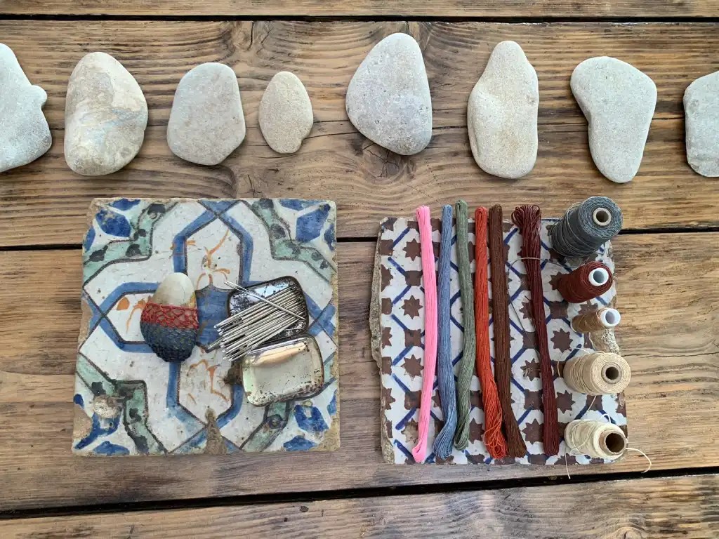 A flat-lay image showcasing various smooth stones arranged in a row on a wooden surface, accompanied by spools of thread, needles, and intricately patterned fabric pieces used for crafting.