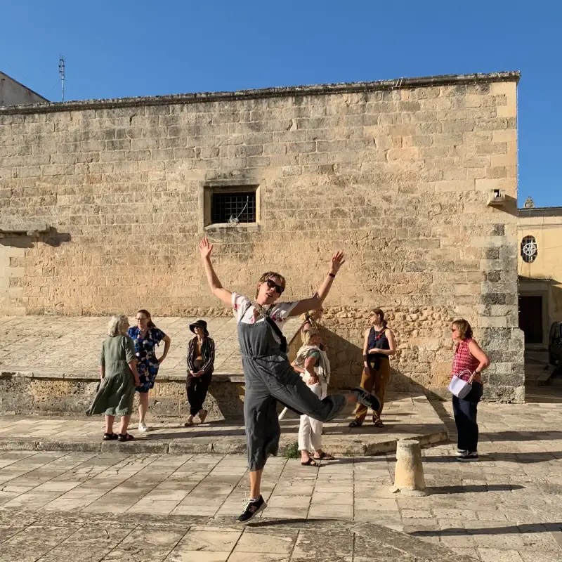 A lively group of people gathered in a sunlit piazza with a stone wall behind them, one person joyfully leaping in the foreground while others observe.