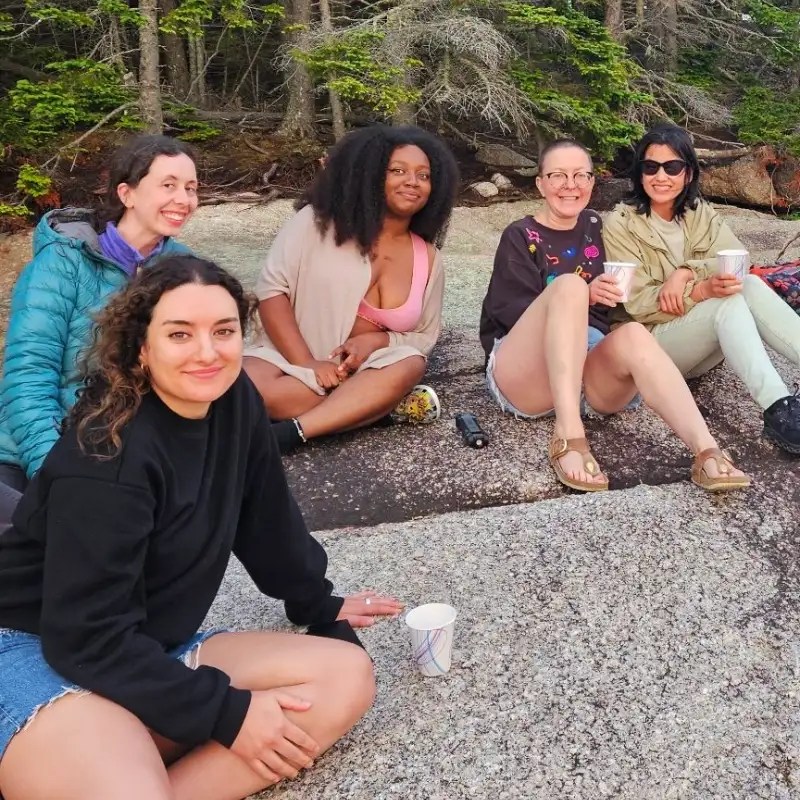A group of five women sitting on rocks in a natural setting, enjoying drinks and smiling at the camera. The background features trees and foliage typical of a coastal landscape.