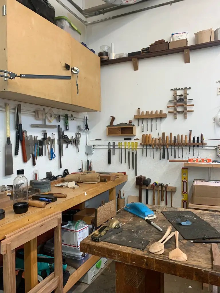 A woodworking studio with various tools organized on the walls and a wooden workbench in the foreground featuring several woodworking projects and tools.