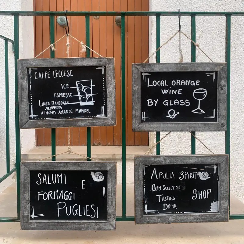Chalkboard signs displaying menu items and offerings, including Caffè Leccese (ice espresso), local orange wine by glass, and selections of Puglian salumi and formaggi; displayed on a green gate.