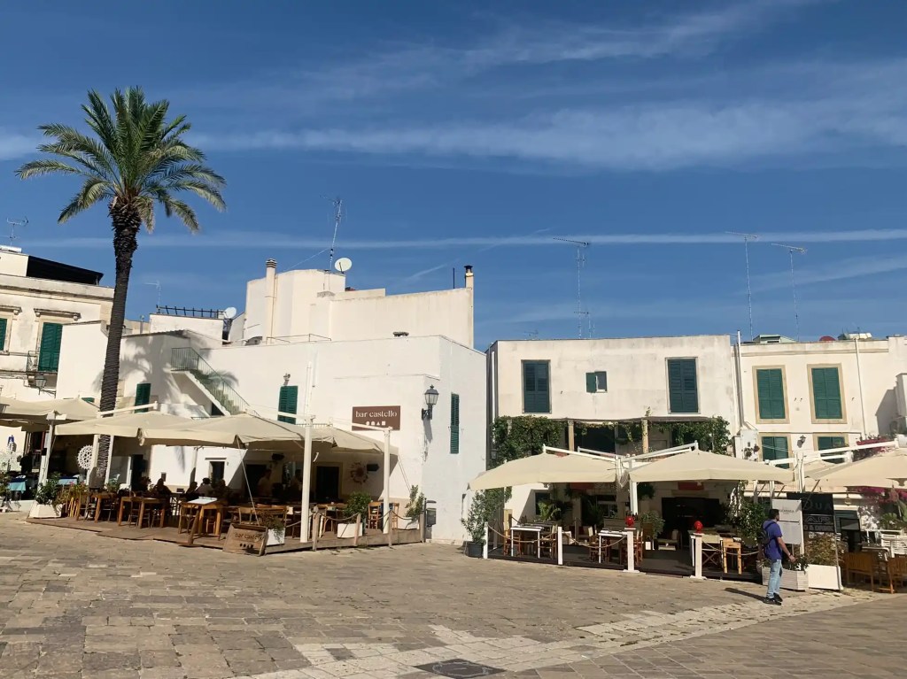 A sunny square in Otranto, Italy, featuring white buildings with green shutters and outdoor dining areas under canopies, with a palm tree in the foreground.