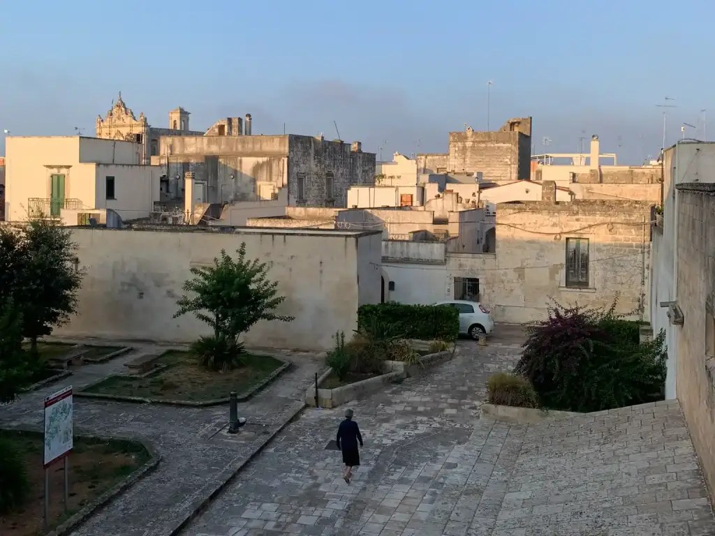 A scenic view of the rooftops and quiet streets in Muro Leccese, Italy, with a lone figure walking down the cobblestone path surrounded by lush greenery and traditional architecture.