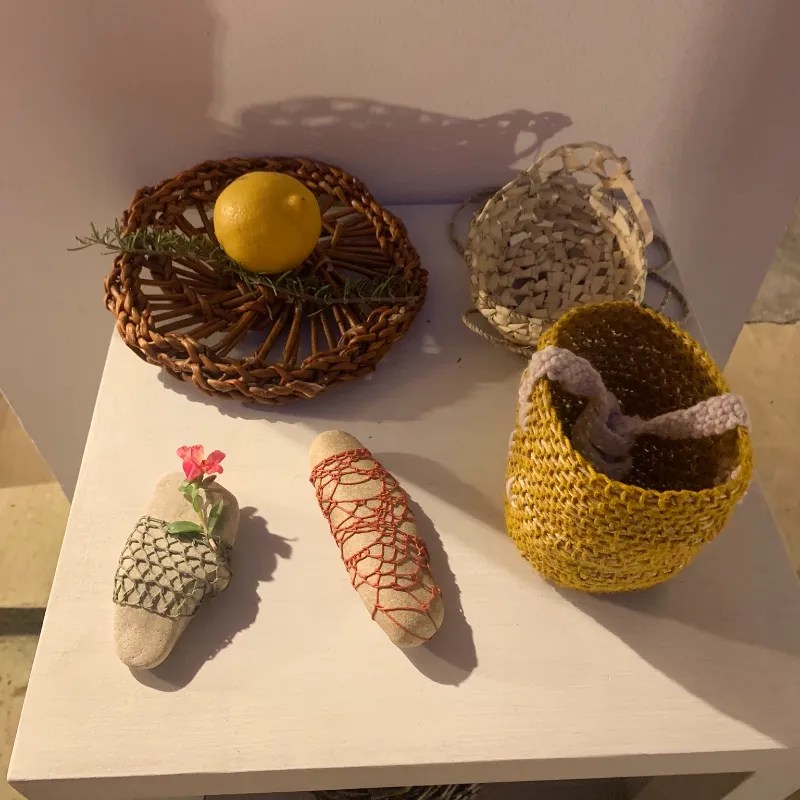 A collection of handmade crafts displayed on a white surface, featuring a woven basket, a decorative plate with a lemon and rosemary, and stone pieces wrapped in colorful twine.