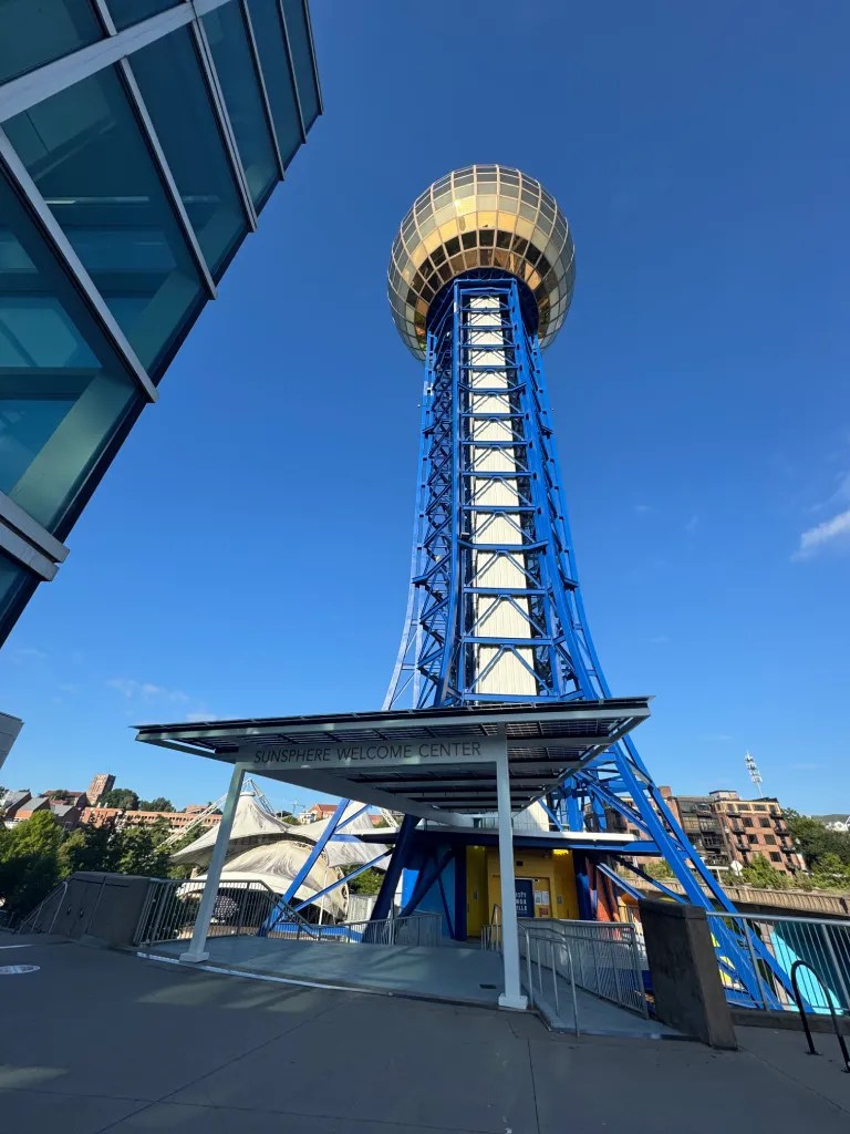 The Sunsphere, a prominent landmark in Knoxville, Tennessee, featuring a golden orb atop a blue steel tower, with the Sunsphere Welcome Center in the foreground and a clear blue sky above.