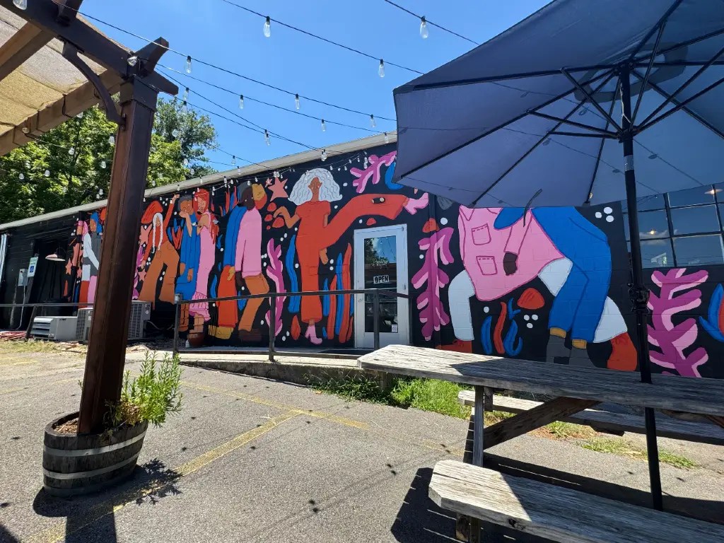 A vibrant mural depicting diverse characters painted on the side of a building, with outdoor seating visible in the foreground under a blue sky.