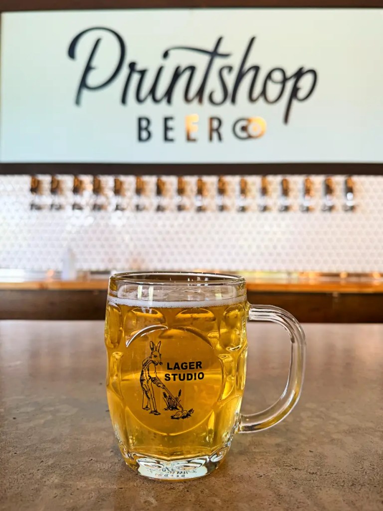 A glass of beer labeled 'LAGER STUDIO' placed on a bar countertop, with a backdrop featuring the Printshop Beer logo.
