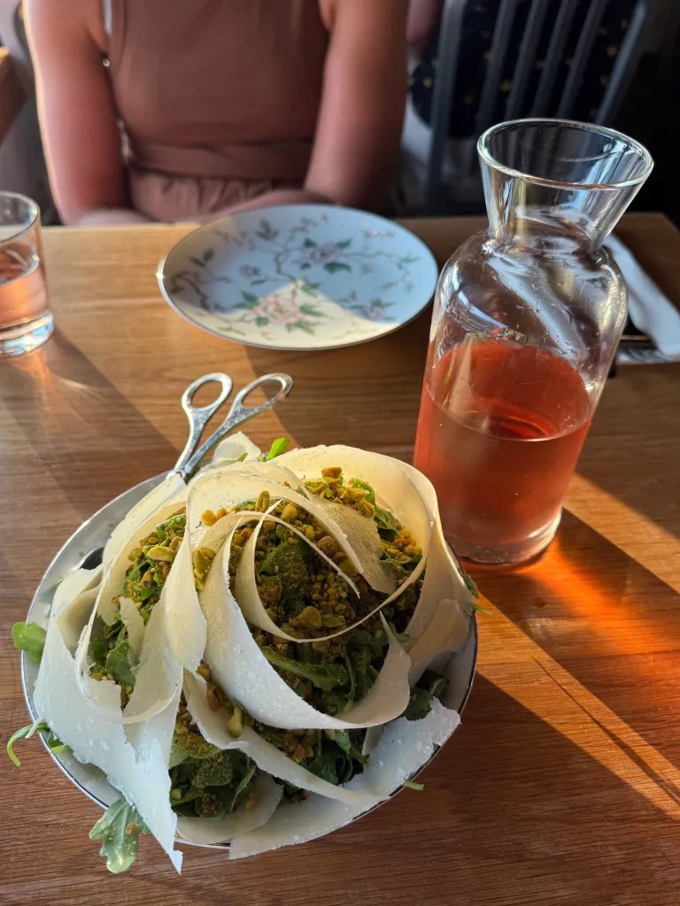 A plate of fresh salad with layers of shaved cheese and pistachios, accompanied by a glass carafe of rosé wine and a decorative floral plate in the background.