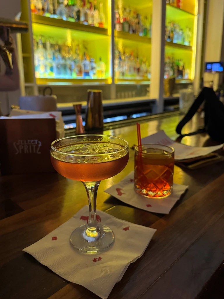 A close-up view of two cocktails on a bar countertop, with a vibrant backdrop of illuminated liquor bottles. The first cocktail, served in a coupe glass, has a light amber color, and the second cocktail is in an old-fashioned glass with a dark amber appearance, each resting on a napkin.