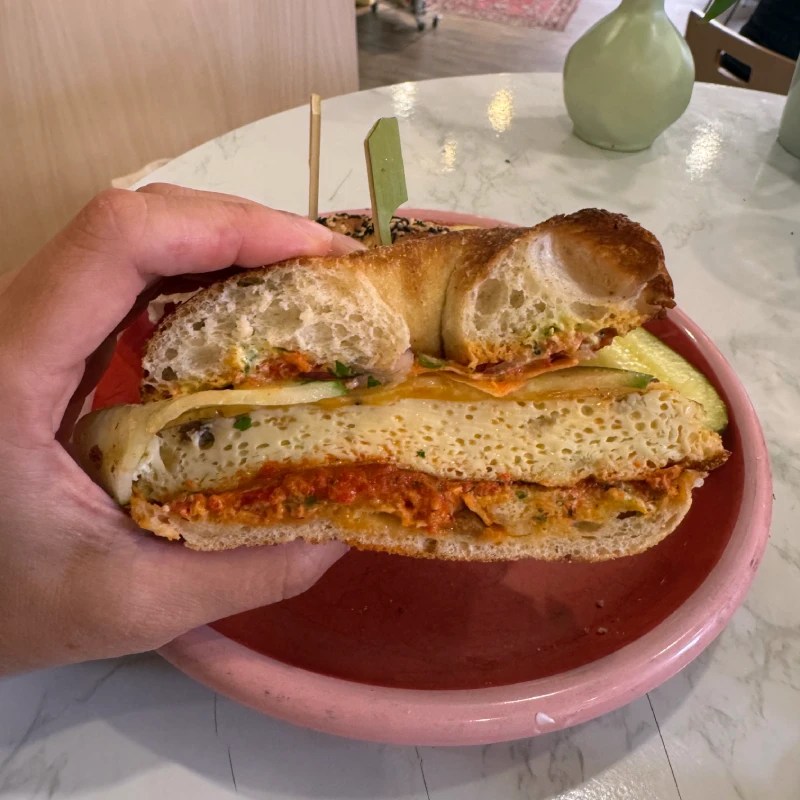 A hand holding a bagel sandwich filled with cheese and and egg frittata, resting on a pink plate.