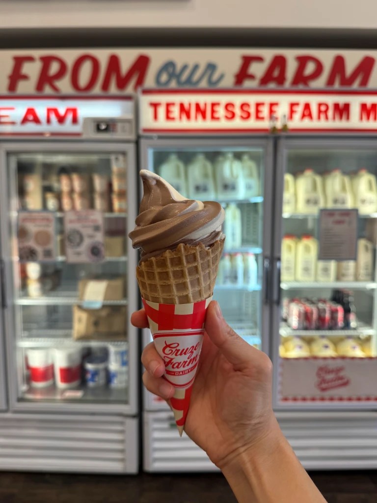A hand holding a waffle cone filled with chocolate swirl ice cream in front of a freezer display at Cruze Farm Dairy, which features various dairy products.