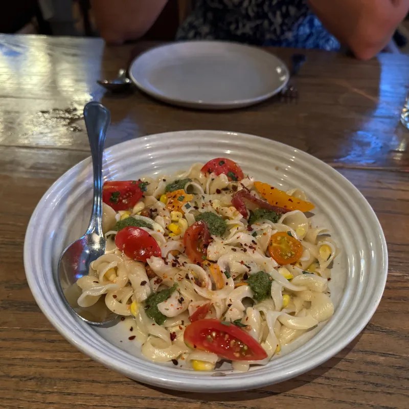 A dish of pasta featuring cherry tomatoes, corn, and greens, served in a white bowl on a wooden table.