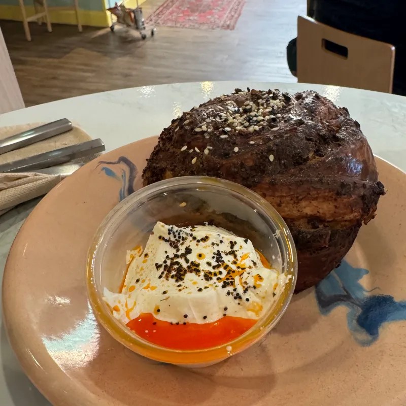 A close-up of a large, round, savory pastry with a crusty top sprinkled with sesame seeds, sitting on a decorative plate alongside a small bowl of creamy dip topped with spices and a drizzle of orange sauce.