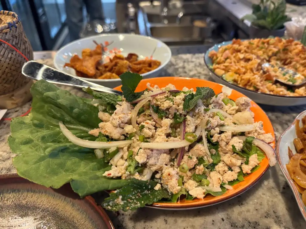 A plate of Laotian chicken salad made with ground chicken, herbs, and spices, served over a bed of leafy greens. In the background, various other dishes can be seen.