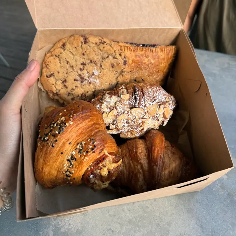 A hand holding a takeout box filled with an assortment of baked goods, including a chocolate chip cookie, pastries, and croissants.