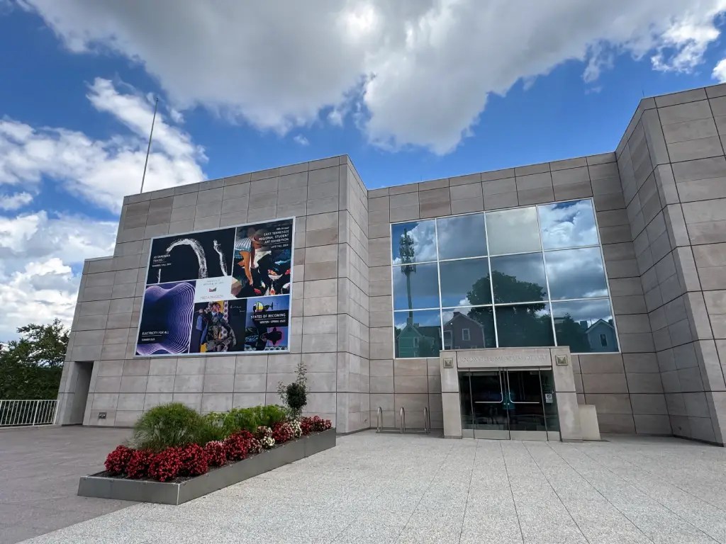 Exterior view of the Knoxville Museum of Art featuring a modern building design with large glass windows and vibrant flower beds in the foreground under a partly cloudy sky.