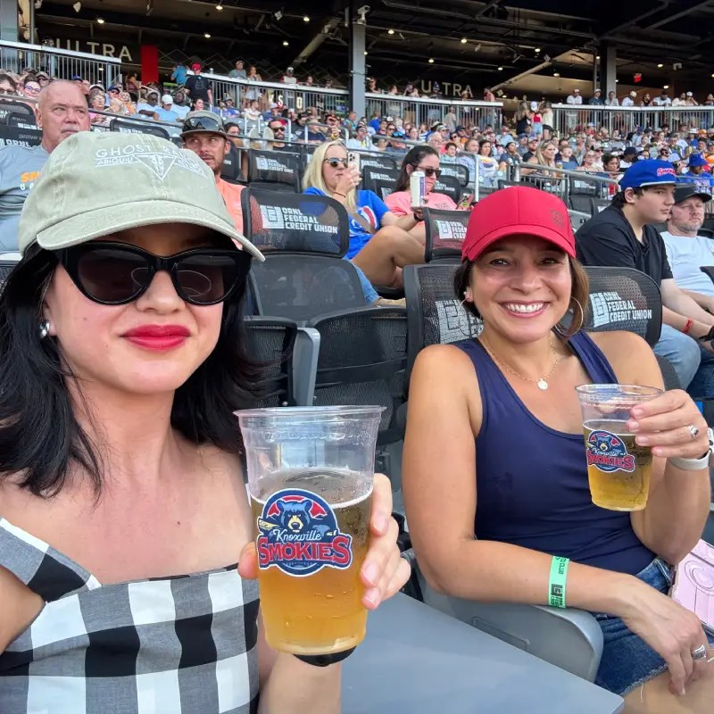 Two women at a baseball game holding cups of beer, smiling at the camera. One woman wears a black and white checkered dress with a cap and sunglasses, while the other wears a red baseball cap and a navy blue tank top. The stadium is filled with spectators in the background.