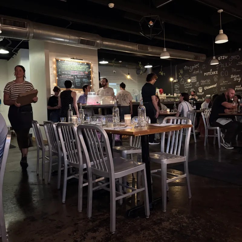 Interior of a bustling restaurant with patrons seated at tables, staff interacting with customers, and a chalkboard wall displaying the menu.