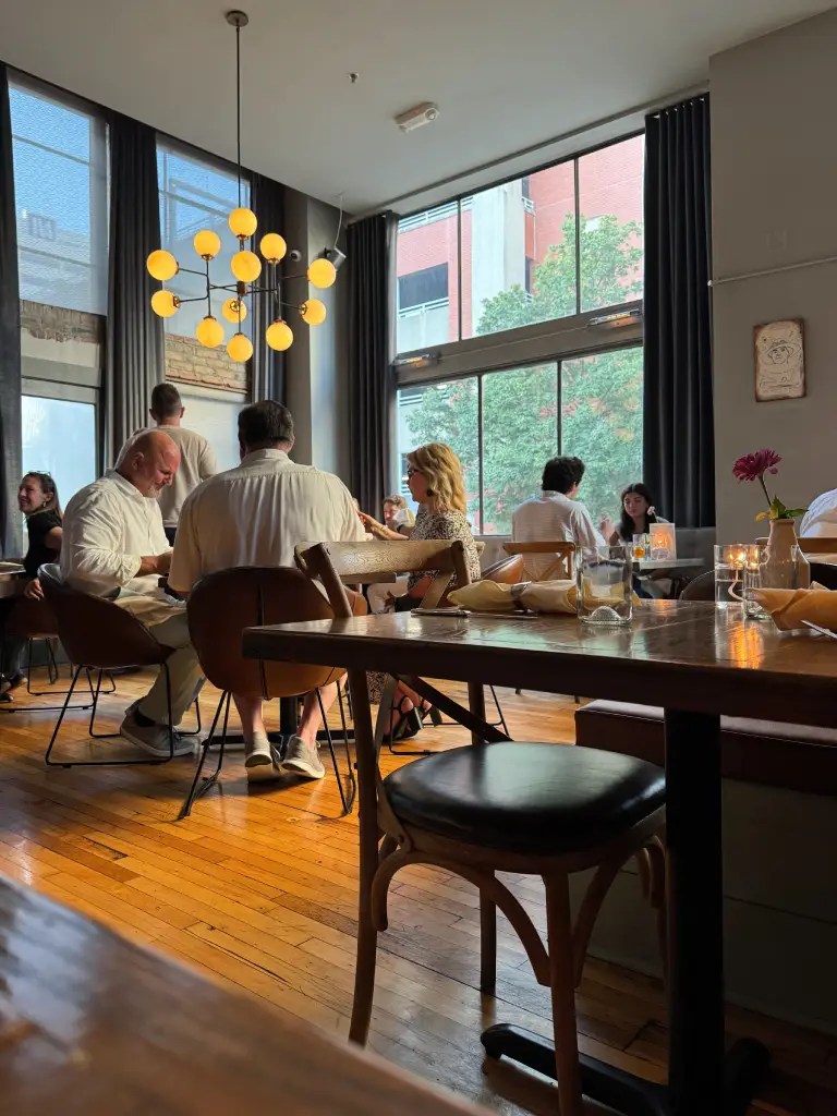 A contemporary dining interior featuring wooden floors, modern furniture, and large windows allowing natural light. Diners are engaged in conversation at various tables under stylish lighting.