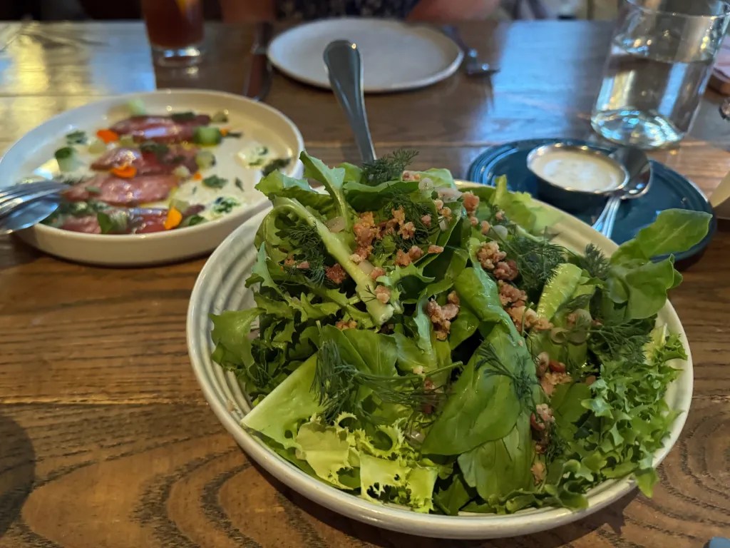 A fresh green salad with assorted leafy greens and herbs, topped with crispy bits of bacon on a wooden table, with a plate of tuna crudo in the background.