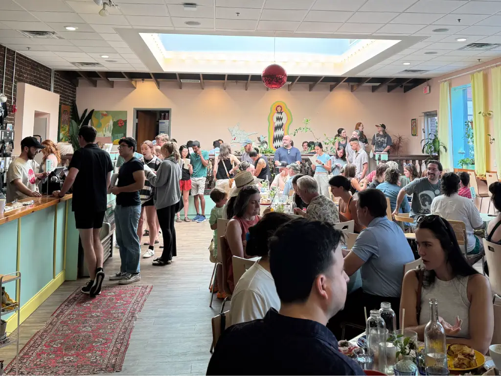 A busy indoor dining area at a restaurant, filled with people enjoying their meals and chatting, with a bar counter in the foreground and colorful decor on the walls.