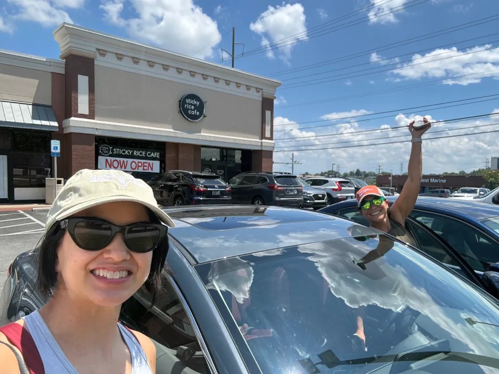 Two people smiling in front of Sticky Rice Cafe, a restaurant with a sign indicating it's now open. They are standing next to a car on a sunny day with blue skies.