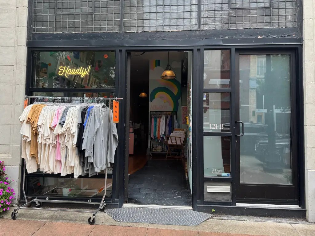 Exterior view of a boutique store featuring a 'Howdy!' neon sign above the entrance, with a clothing rack displaying various shirts and hoodies in front of the shop.