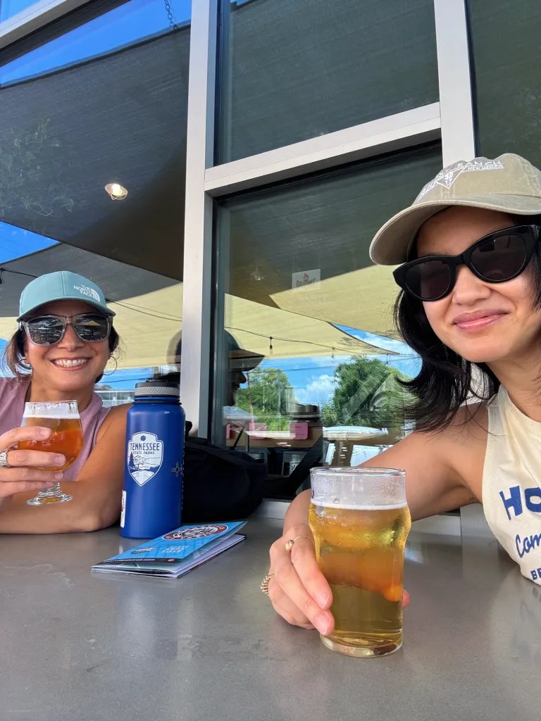 Two women enjoying drinks at a bar, one holding a glass of light beer and the other a glass of a West Coast IPA. They are seated at an outdoor table with a water bottle and a menu in front of them. The setting appears bright and relaxed.
