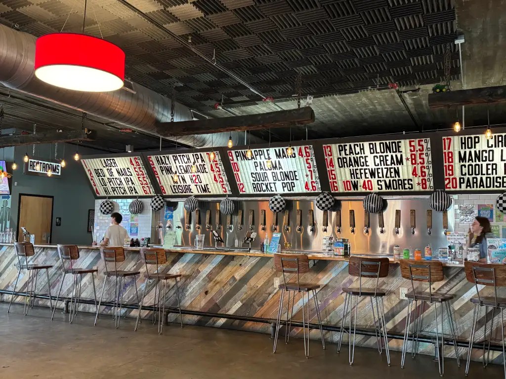 Interior view of a brewery bar featuring a wooden counter, high stools, and a menu board displaying various beer options and their prices.