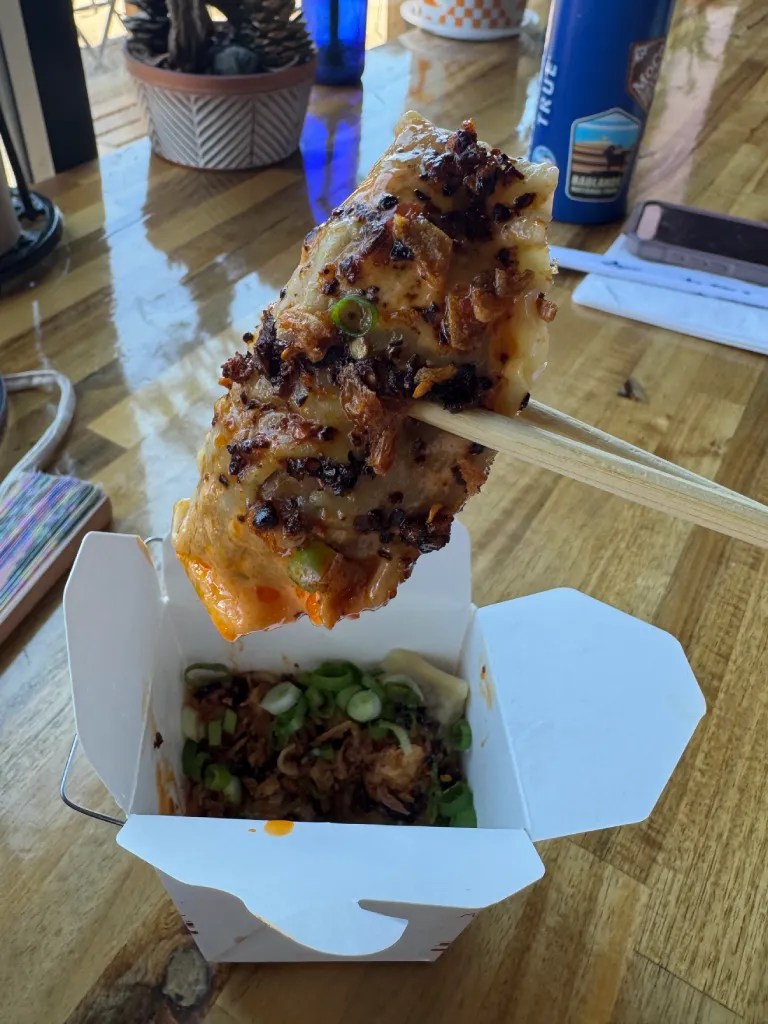 A close-up of a crispy dumpling held with chopsticks, revealing layers of browned dough, garnished with green onions, sitting above a takeout container filled with fried shallots and scallions.