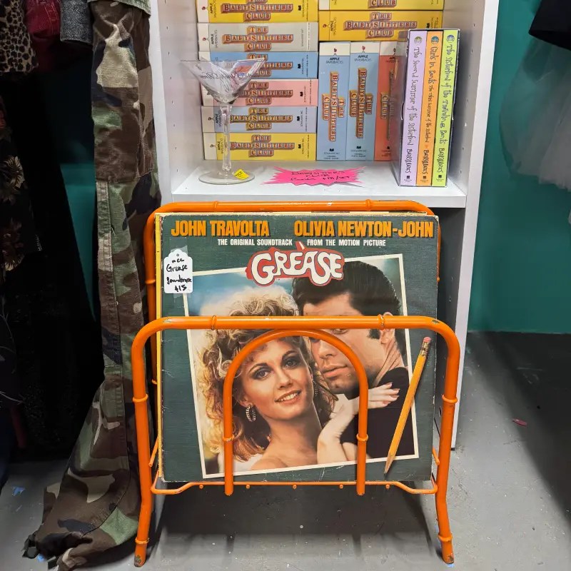 A vintage record display featuring the soundtrack of 'Grease' by John Travolta and Olivia Newton-John, framed in an orange display stand next to a glass and a collection of The Babysitters Club books.