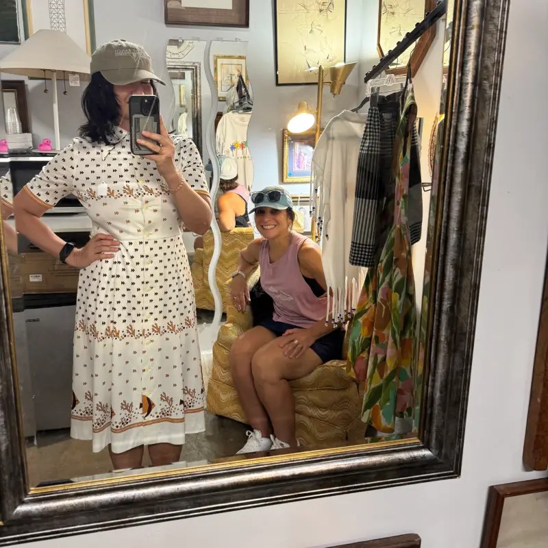 A woman stands in front of a mirror, taking a selfie in a white patterned dress while a friend sits on a couch in the background, both in a cozy boutique filled with clothing and accessories.