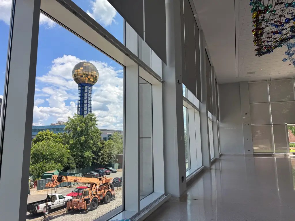 A view from inside the Knoxville Museum of Art showcasing large windows looking out to a cityscape featuring the Sunsphere, a prominent gold-globe structure, surrounded by trees and construction vehicles.