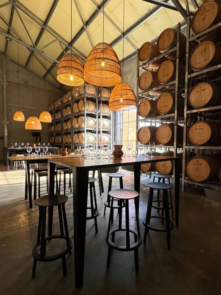 Interior of Cherubino winery showcasing a tasting area with a large table and stools, surrounded by oak wine barrels, and illuminated by woven pendant lights.