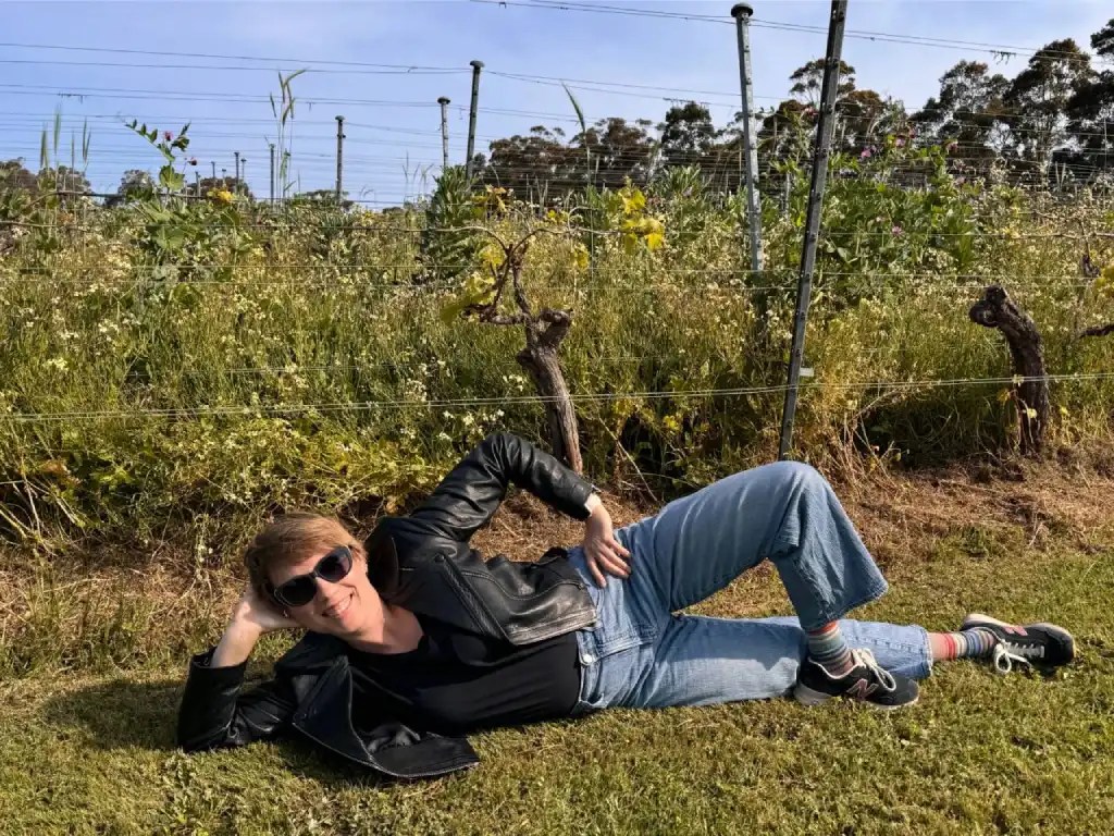 A woman lounging in a vineyard, wearing sunglasses, a leather jacket, and casual blue jeans, surrounded by wildflowers and grapevines under a clear sky.