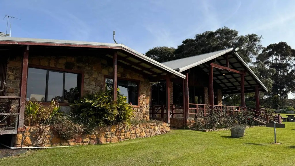 Exterior view of the Cullen Winery tasting room from the garden: a charming stone and wooden house with a large porch, surrounded by greenery and trees, located in a serene outdoor setting.