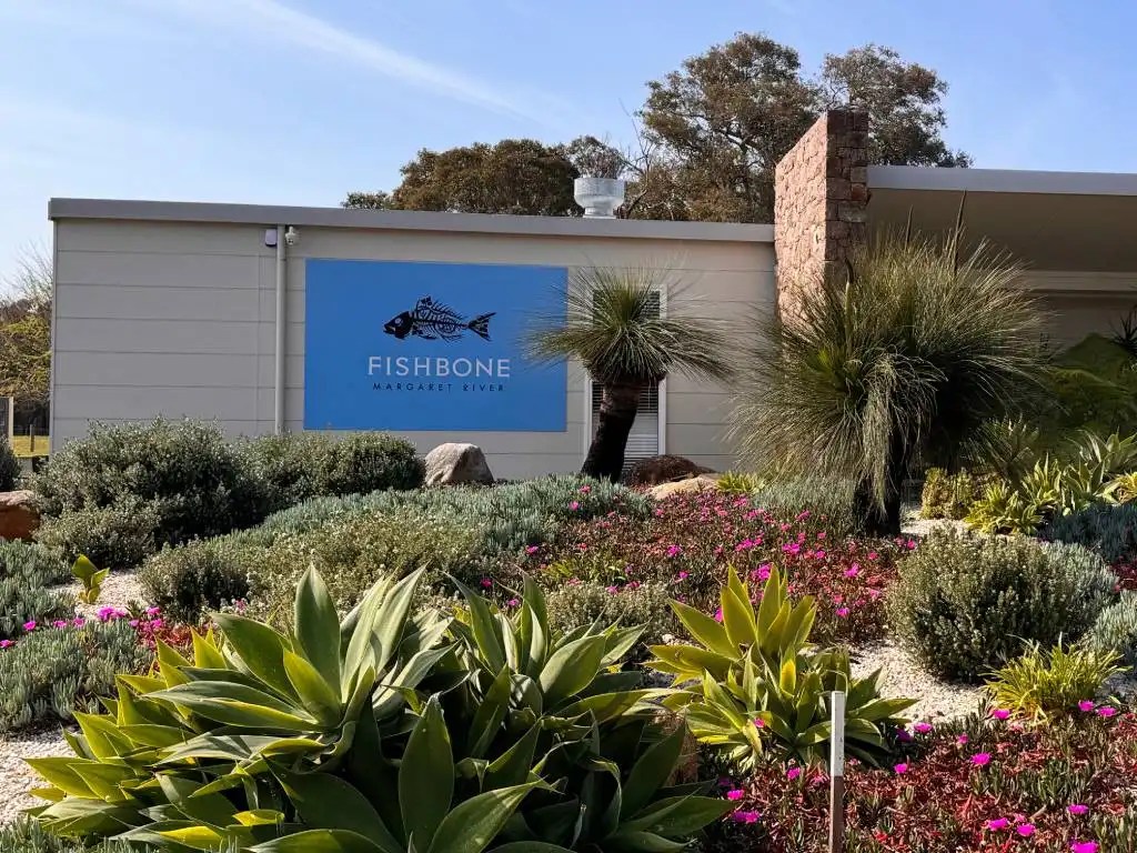 Exterior view of Fishbone Winery, showcasing a blue sign with the name and artwork of a fish, surrounded by lush landscaping with various plants and flowers.
