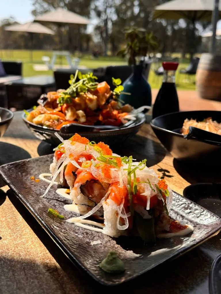 A beautifully arranged platter featuring sushi rolls topped with salmon, avocado, and green onions, accompanied by stylish side dishes, set against a sunny outdoor dining area.